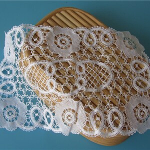 以下が含まれることがあります： 木製のパンバスケットの上に置かれた、繊細な円形と楕円形の模様が施された白いレースのドイリーです。