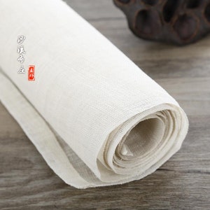 Puede incluir: Un rollo de tela blanca, posiblemente lino o algod&oacute;n, est&aacute; enrollado apretadamente. La tela es lisa y tiene una textura sutil. El rollo est&aacute; sobre una superficie de madera.