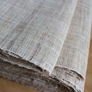 May include: A close-up of a beige woven fabric, folded and rolled up. The fabric has a natural, textured look and feel.