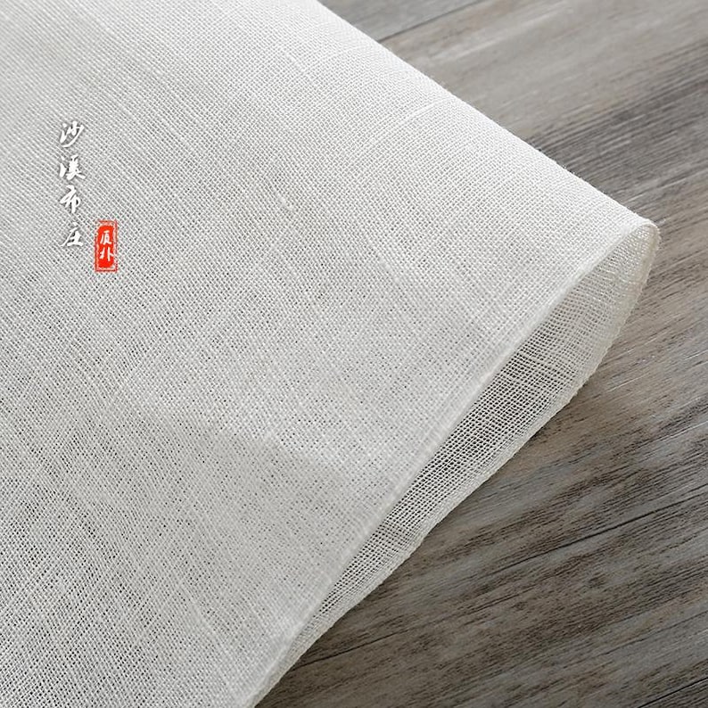 Puede incluir: Tela de gasa blanca con un tejido fino, doblada sobre una superficie de madera. La tela tiene el texto "纱 质 布" en caracteres negros y un sello rojo.