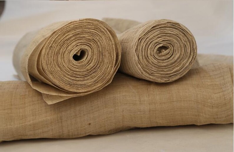 May include: Two rolls of natural beige fabric, likely burlap or linen, are rolled up and placed on a flat surface. The fabric is woven and has a slightly rough texture.