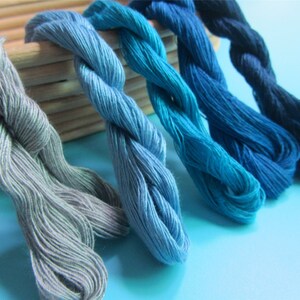 May include: A selection of embroidery threads in various shades of blue and grey. The colours range from pale grey to a deep navy. The threads are twisted and appear soft, with a wooden box in the background. The image is well-lit.