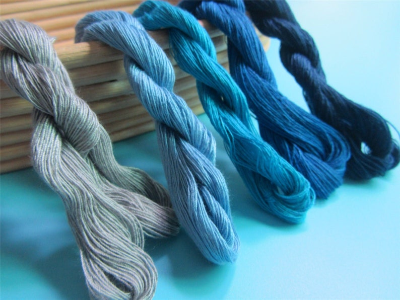 May include: A selection of embroidery threads in various shades of blue and grey. The colours range from pale grey to a deep navy. The threads are twisted and appear soft, with a wooden box in the background. The image is well-lit.
