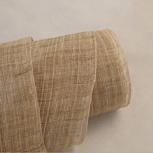May include: A roll of natural brown burlap fabric with a woven texture. The fabric is rolled up and the edges are visible.