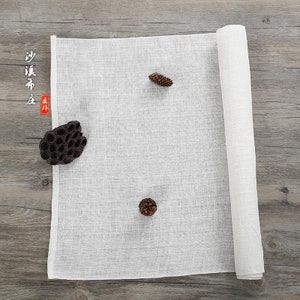 Puede incluir: Un rollo de tela de algod&oacute;n blanco, desenrollado y colocado plano sobre una superficie de madera. La tela es lisa y tiene una textura sutil. Dos peque&ntilde;as pi&ntilde;as y una c&aacute;psula de loto seca se colocan sobre la tela.