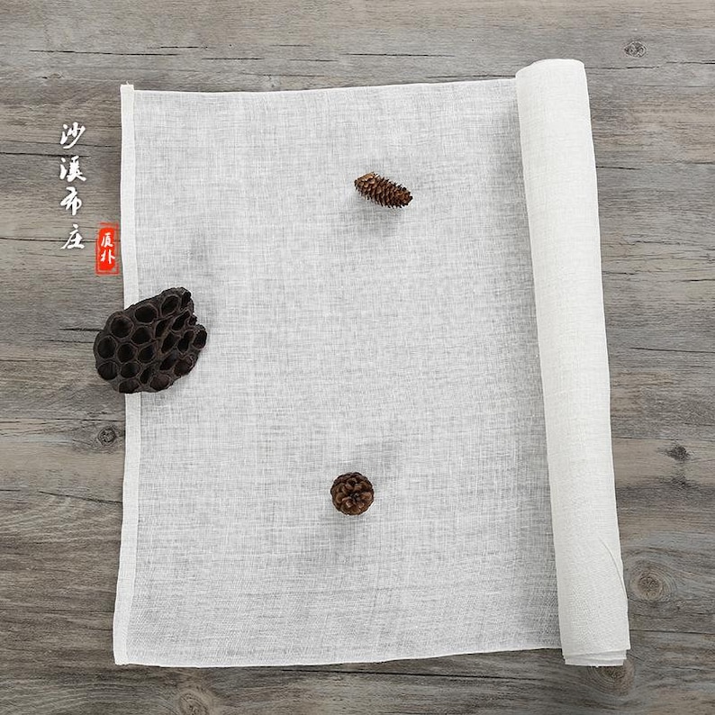 Puede incluir: Un rollo de tela de algod&oacute;n blanco, desenrollado y colocado plano sobre una superficie de madera. La tela es lisa y tiene una textura sutil. Dos peque&ntilde;as pi&ntilde;as y una c&aacute;psula de loto seca se colocan sobre la tela.