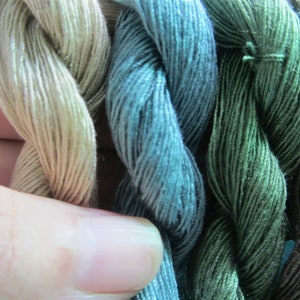 May include: A close-up of several skeins of embroidery thread in shades of green, blue, and tan. The threads are tightly twisted, revealing their texture. The colours vary from light, almost white, to deep forest green and dark brown.