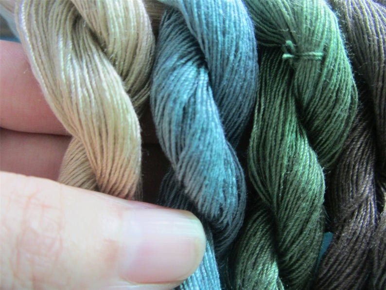 May include: A close-up of several skeins of embroidery thread in shades of green, blue, and tan. The threads are tightly twisted, revealing their texture. The colours vary from light, almost white, to deep forest green and dark brown.