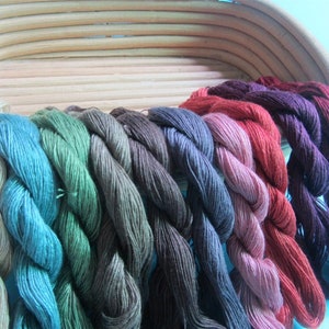 May include: A selection of colourful embroidery floss in various shades, including teal, green, red, and purple. The floss is arranged in a row, with each colour neatly twisted. The background features a light blue wall and a woven basket.