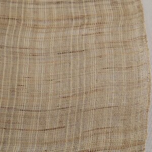 May include: A close-up of a beige woven fabric with a subtle brown and white pattern. The fabric has a rough texture and a slightly uneven weave.