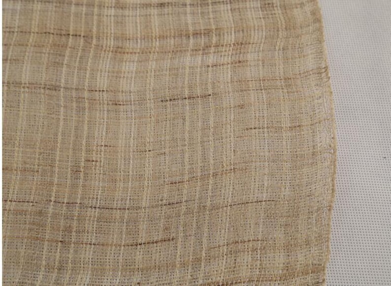May include: A close-up of a beige woven fabric with a subtle brown and white pattern. The fabric has a rough texture and a slightly uneven weave.