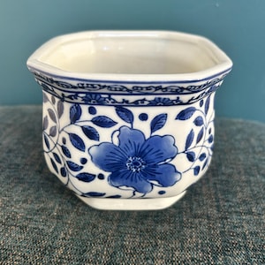 May include: A white ceramic planter with a hexagonal shape, featuring a blue floral pattern and a decorative border around the rim. The exterior displays blue painted flowers and leaves on a white background, with a smooth, glossy finish.