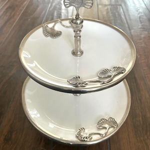 May include: A two-tiered serving stand featuring white circular plates with silver-toned rims. The central metal post is topped with a ginkgo leaf finial, and additional silver-toned ginkgo leaf accents are attached to the surface of each plate.