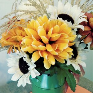May include: A green metal milk can vase filled with a bouquet of yellow, white, and orange artificial flowers. The flowers include sunflowers, mums, and wheat.