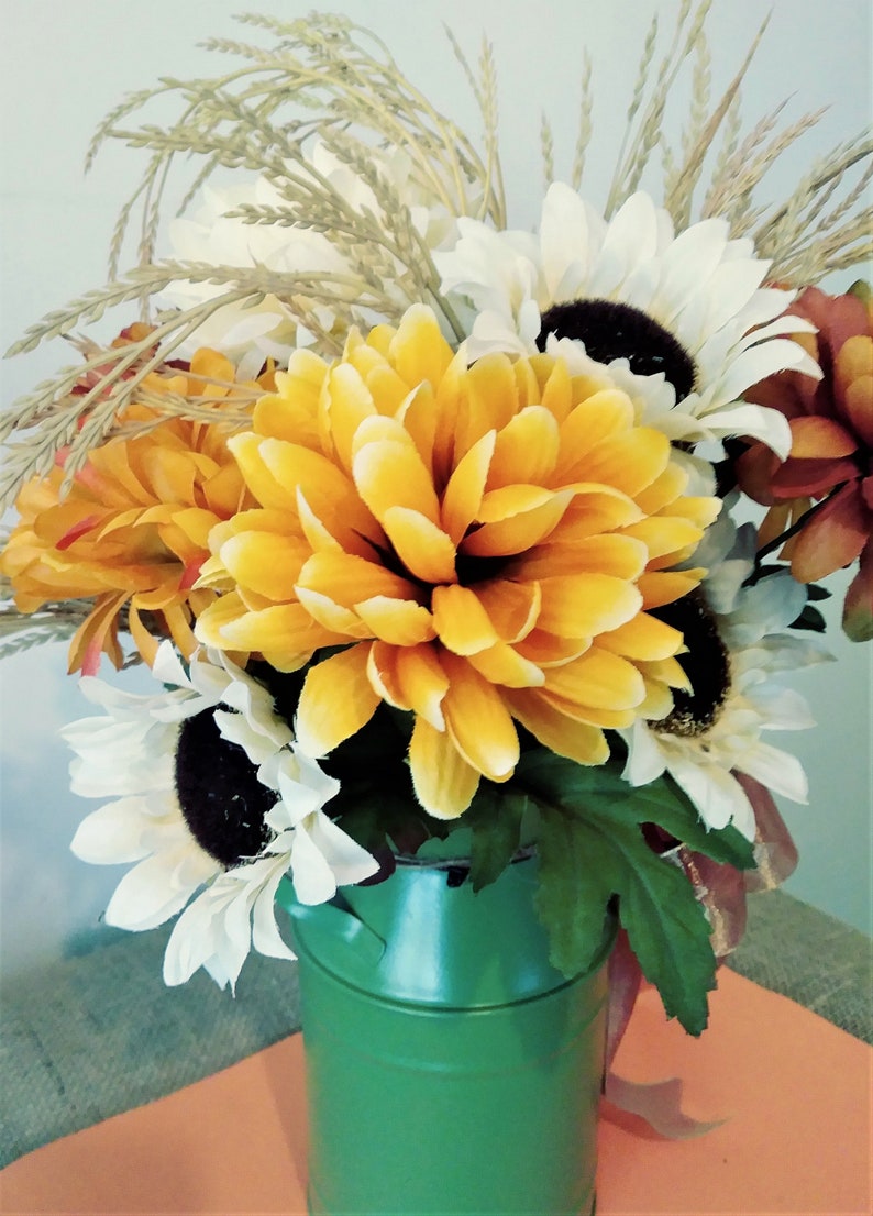 May include: A green metal milk can vase filled with a bouquet of yellow, white, and orange artificial flowers. The flowers include sunflowers, mums, and wheat.