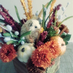 May include: A wooden box filled with autumnal decor, including orange and white pumpkins, a white hedgehog, and orange and burgundy flowers. The box has a wooden leaf with the text "Gather" on it.