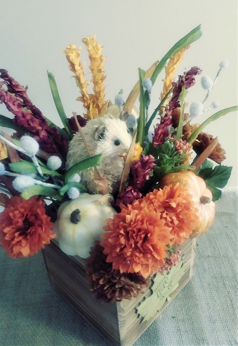 May include: A wooden box filled with autumnal decor, including orange and white pumpkins, a white hedgehog, and orange and burgundy flowers. The box has a wooden leaf with the text "Gather" on it.