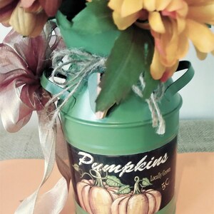 May include: A green metal milk can with a brown burlap bow and a floral arrangement of yellow and red flowers. The can has a sticker that says "Pumpkins" and "Locally Grown 5c".