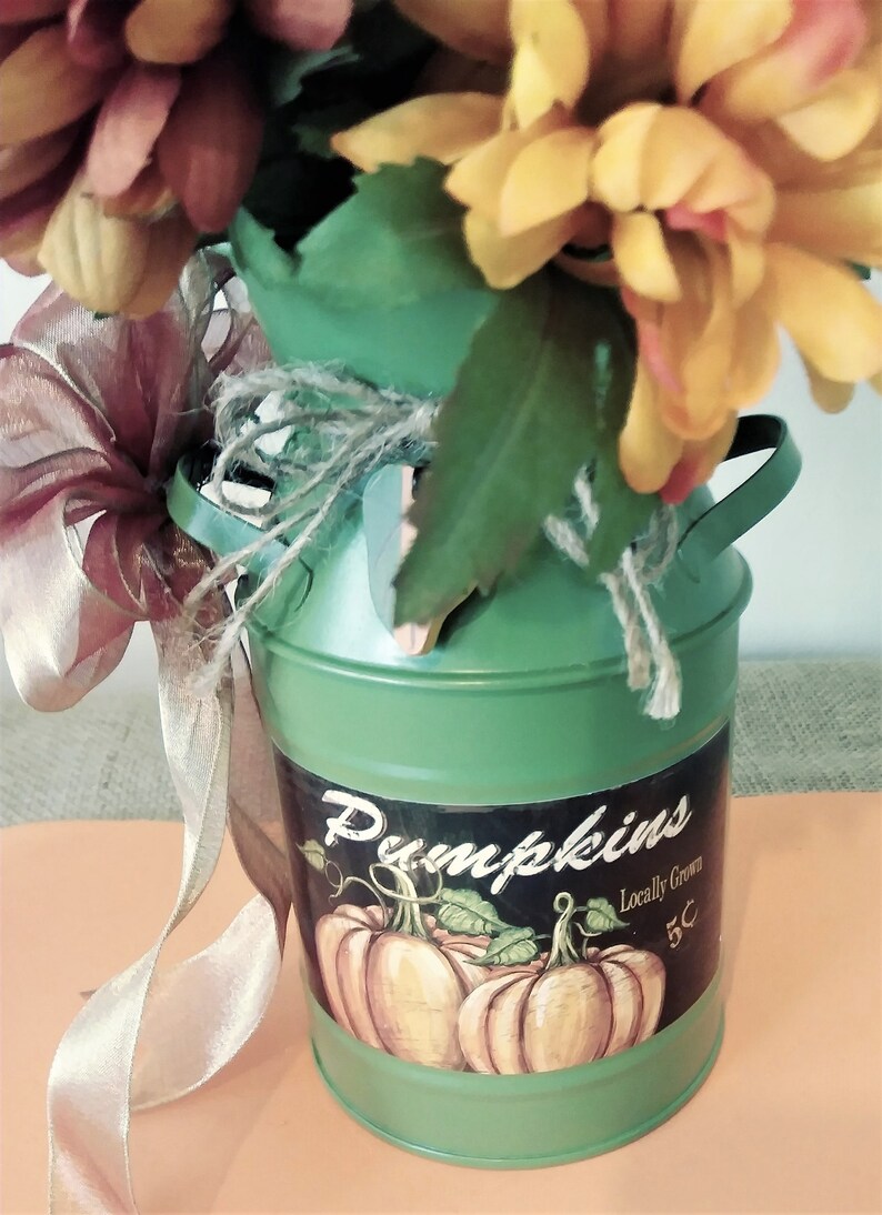 May include: A green metal milk can with a brown burlap bow and a floral arrangement of yellow and red flowers. The can has a sticker that says "Pumpkins" and "Locally Grown 5c".
