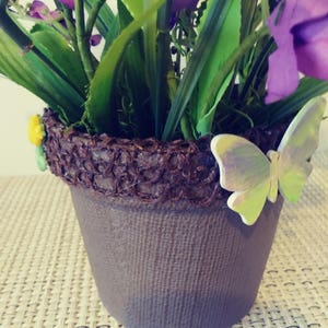 May include: A purple flower arrangement in a brown pot with a decorative brown woven rim. A green butterfly with iridescent wings is attached to the rim of the pot.