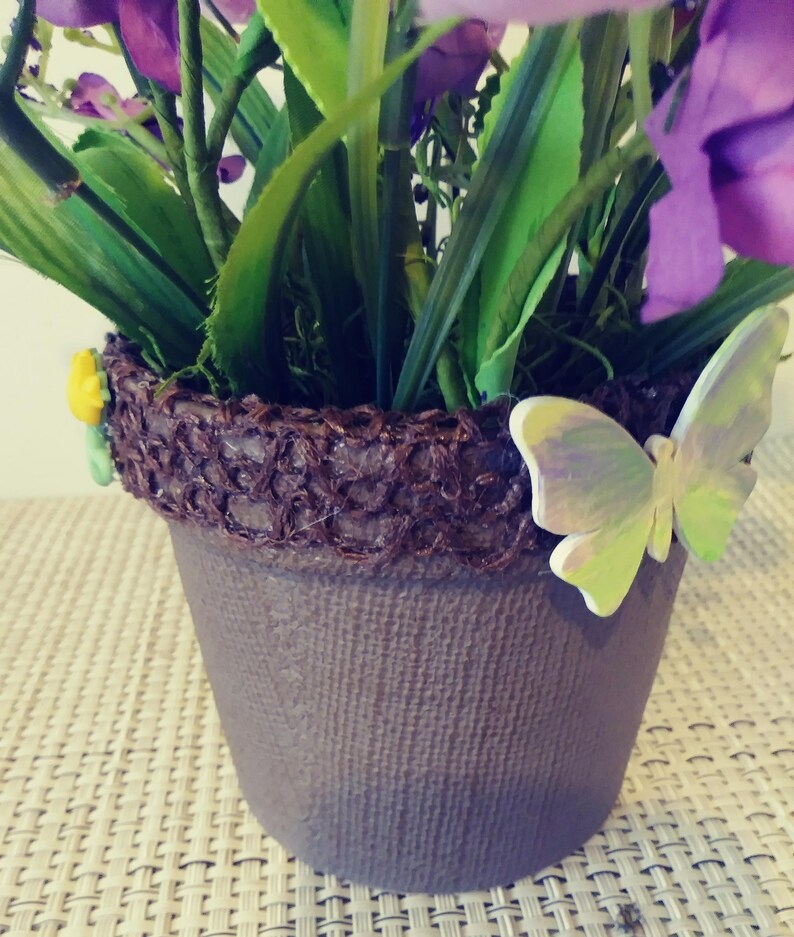 May include: A purple flower arrangement in a brown pot with a decorative brown woven rim. A green butterfly with iridescent wings is attached to the rim of the pot.