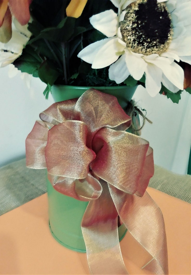May include: A green metal pitcher with a brown and gold ribbon bow. The pitcher is filled with a bouquet of artificial sunflowers.