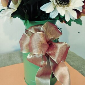 May include: A green metal vase with a brown and gold ribbon bow. The vase is filled with a bouquet of artificial sunflowers.