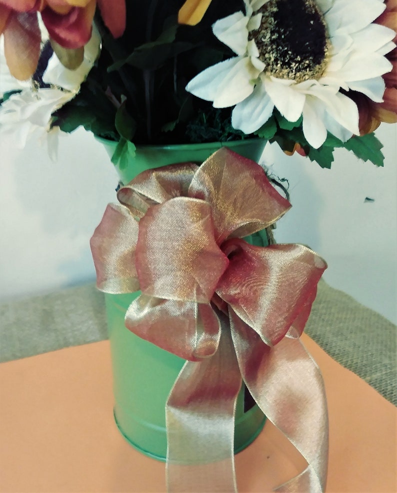 May include: A green metal vase with a brown and gold ribbon bow. The vase is filled with a bouquet of artificial sunflowers.