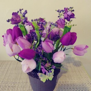 May include: A purple and lavender artificial flower arrangement in a brown pot with a butterfly decoration. The arrangement includes tulips and small purple flowers.