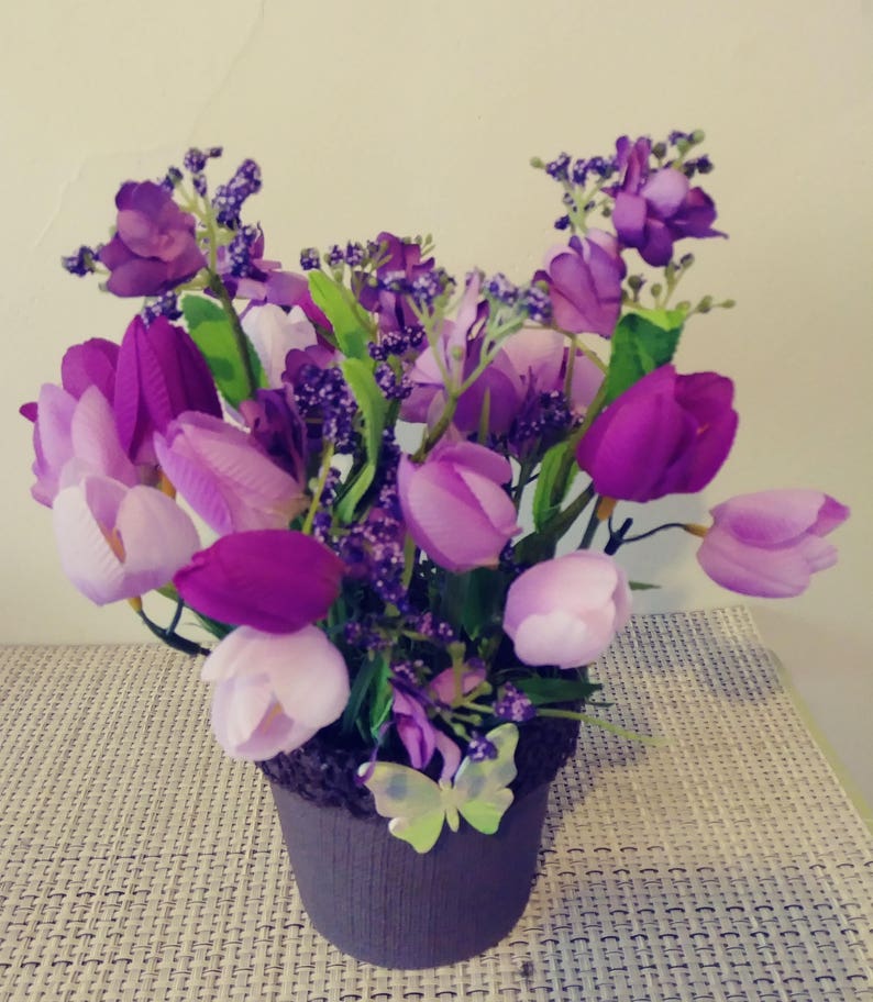 May include: A purple and lavender artificial flower arrangement in a brown pot with a butterfly decoration. The arrangement includes tulips and small purple flowers.