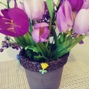 May include: A purple and white flower arrangement in a brown pot with a decorative brown rim. The pot is decorated with small yellow and blue flowers.