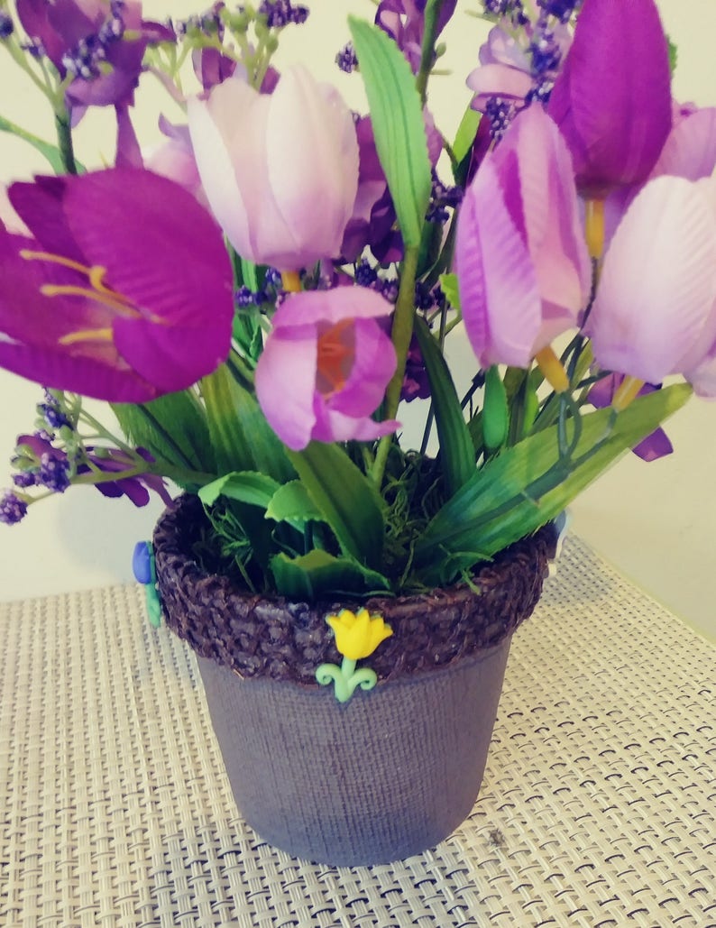 May include: A purple and white flower arrangement in a brown pot with a decorative brown rim. The pot is decorated with small yellow and blue flowers.