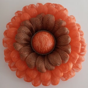 May include: A large orange and brown mesh wreath with a textured orange centre. The wreath is made of multiple layers of mesh and has a rustic, farmhouse style.