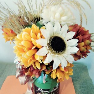 May include: A green metal watering can with a pumpkin design and the text "Penny Lane" is filled with a bouquet of fall flowers. The bouquet includes white, yellow, and orange flowers, as well as dried grasses.