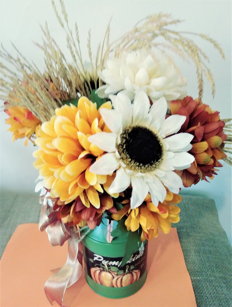 May include: A green metal watering can with a pumpkin design and the text "Penny Lane" is filled with a bouquet of fall flowers. The bouquet includes white, yellow, and orange flowers, as well as dried grasses.