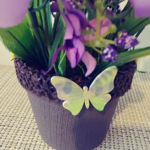 May include: A small brown pot with a decorative brown woven rim, filled with purple flowers and green leaves. A light green butterfly with iridescent wings is attached to the pot.