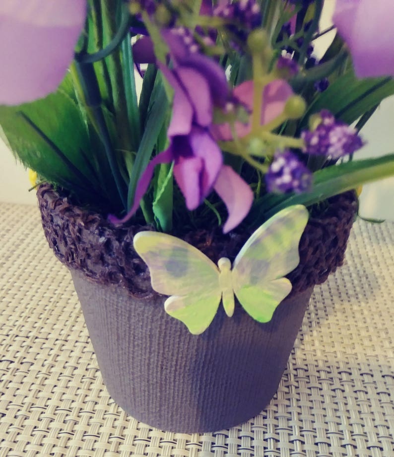 May include: A small brown pot with a decorative brown woven rim, filled with purple flowers and green leaves. A light green butterfly with iridescent wings is attached to the pot.
