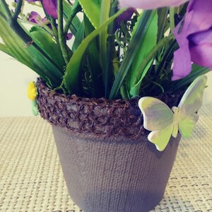 May include: A purple flower arrangement in a brown pot with a burlap-like texture. The pot is decorated with a brown woven band and a green and purple butterfly.