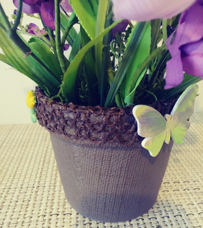 May include: A purple flower arrangement in a brown pot with a burlap-like texture. The pot is decorated with a brown woven band and a green and purple butterfly.