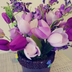 May include: A purple and white flower arrangement in a brown pot with a textured rim. The flowers are in a variety of shades of purple and white, and the pot is sitting on a white wicker surface.