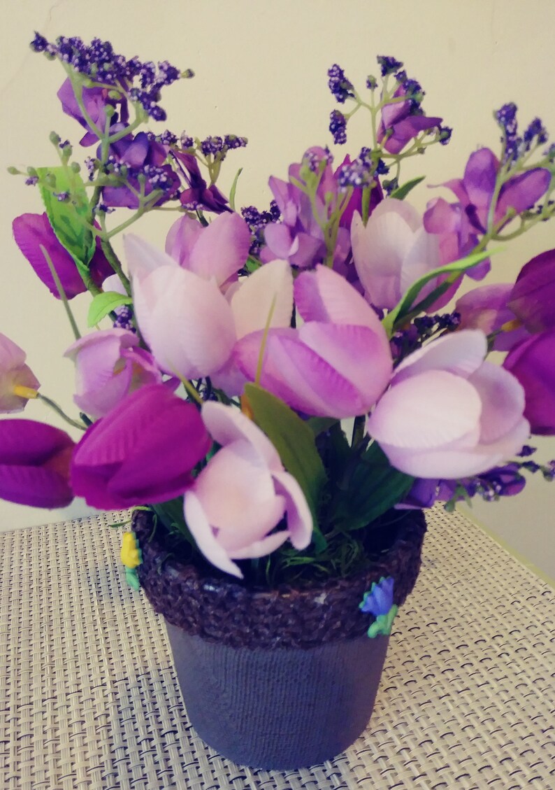 May include: A purple and white flower arrangement in a brown pot with a textured rim. The flowers are in a variety of shades of purple and white, and the pot is sitting on a white wicker surface.