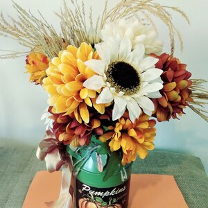 May include: A green metal milk can with a fall-themed design featuring pumpkins and the text "Pumpkins Locally Grown 5&cent;" is filled with a bouquet of fall flowers. The bouquet includes white, yellow, and orange chrysanthemums, a white sunflower, and dried wheat stalks.