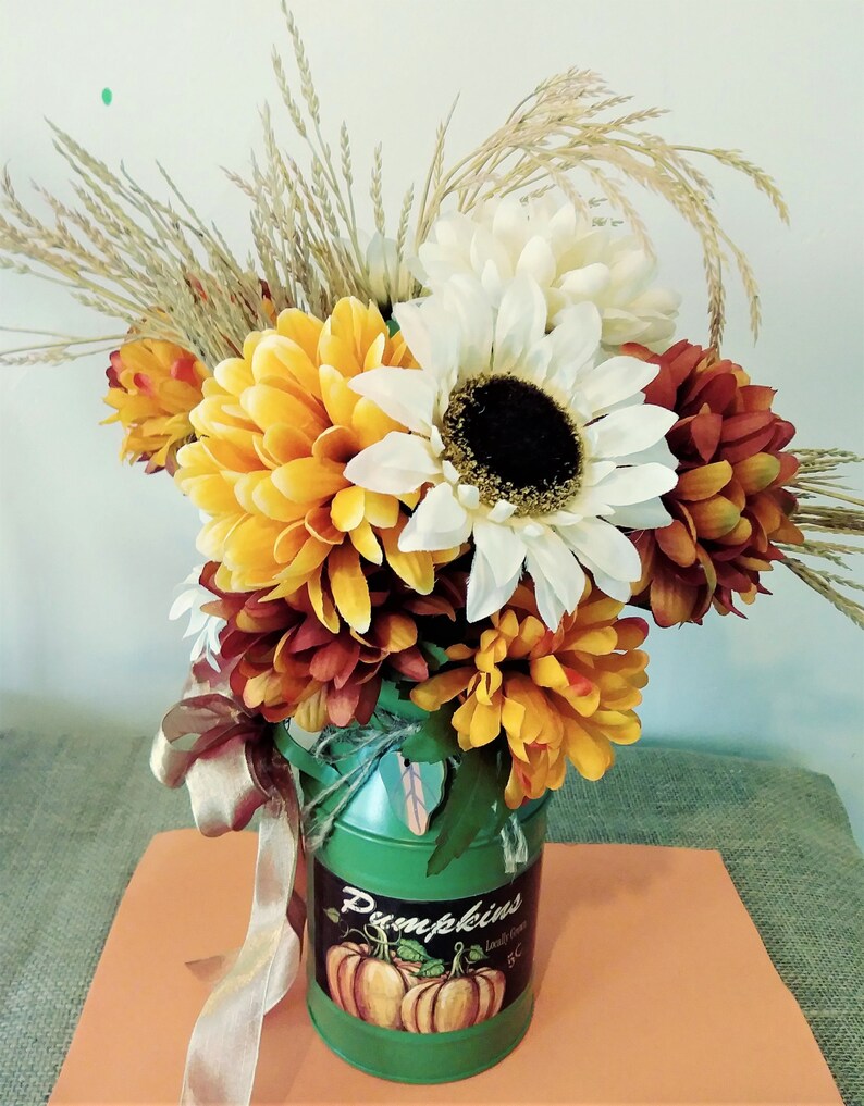 May include: A green metal milk can with a fall-themed design featuring pumpkins and the text "Pumpkins Locally Grown 5&cent;" is filled with a bouquet of fall flowers. The bouquet includes white, yellow, and orange chrysanthemums, a white sunflower, and dried wheat stalks.