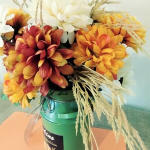 May include: A green milk jug with a label that says "Pumpkins" is filled with a fall floral arrangement. The arrangement includes white, orange, and red flowers, as well as dried wheat stalks.