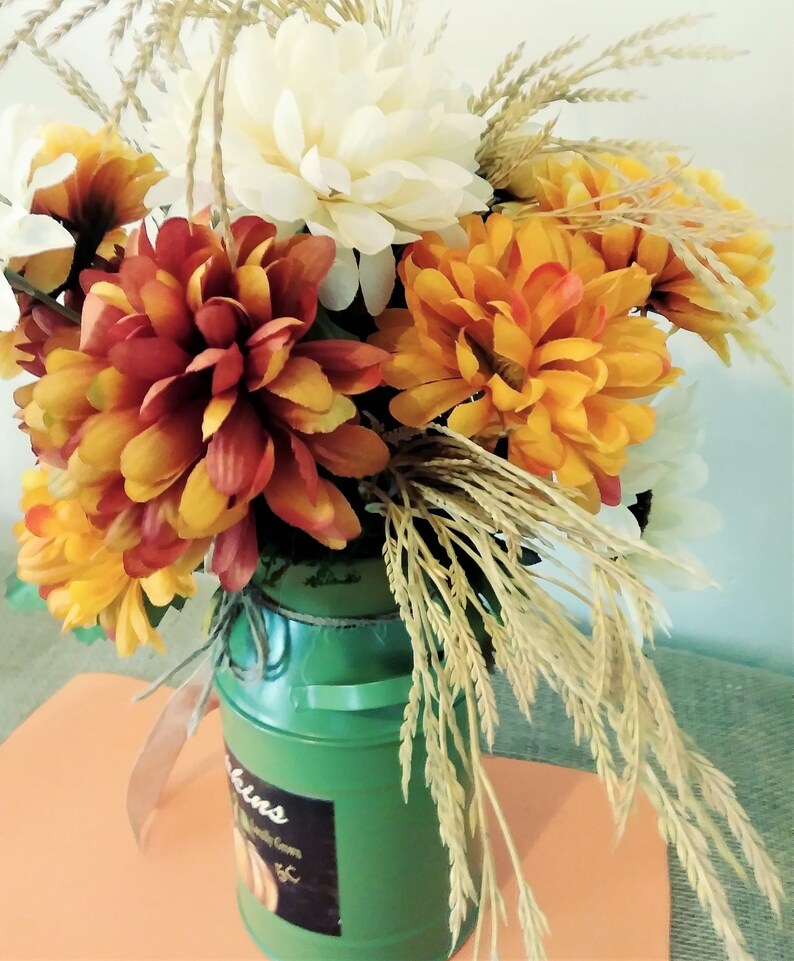 May include: A green milk jug with a label that says "Pumpkins" is filled with a fall floral arrangement. The arrangement includes white, orange, and red flowers, as well as dried wheat stalks.
