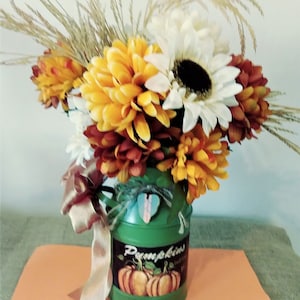 May include: A green metal milk can vase with a pumpkin design and the word "Pumpkins" on the side. The vase is filled with a bouquet of yellow, orange, and white flowers, including sunflowers and mums. A brown ribbon is tied around the handle of the vase.