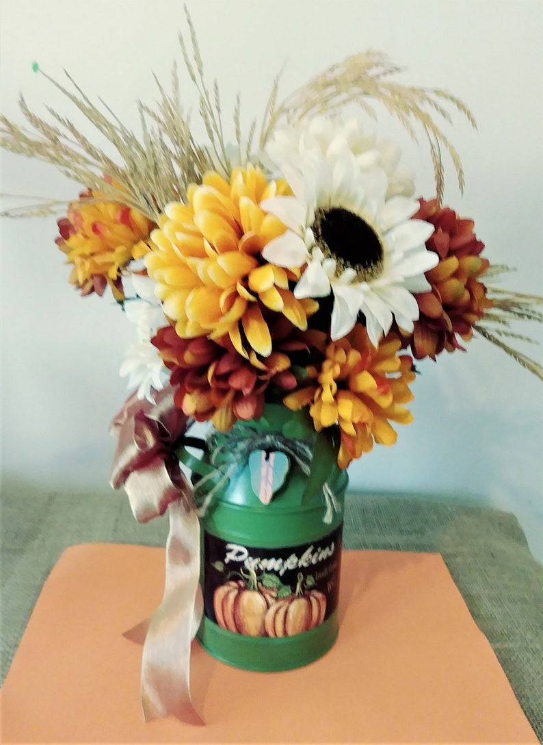 May include: A green metal milk can vase with a pumpkin design and the word "Pumpkins" on the side. The vase is filled with a bouquet of yellow, orange, and white flowers, including sunflowers and mums. A brown ribbon is tied around the handle of the vase.