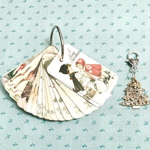 Op de afbeelding: Een verzameling vintage kerstthema-tags uitgespreid op een lichtblauwe achtergrond. De tags worden bij elkaar gehouden door een zilveren ring. Een bedel met een met strass bezette kerstboom hangt naast de tags.