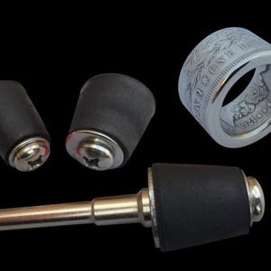 May include: A collection of items on a black background, including two black rubber stoppers with silver screws, a silver ring made from a coin, and a wine bottle stopper with a silver stem and black rubber top. The ring has detailed engravings.
