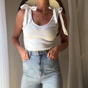 May include: A white ribbed tank top with tie straps and a V-neck is paired with light blue denim jeans. The jeans have a high waist and a relaxed fit. The outfit is simple and casual, suitable for everyday wear.